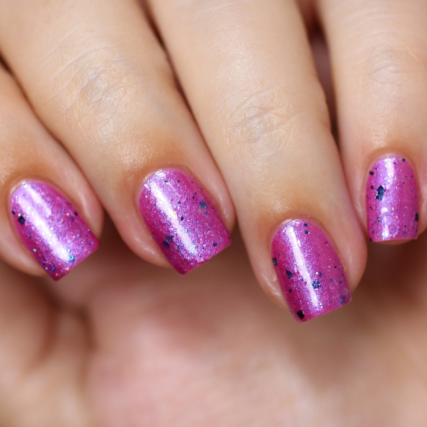 Close-up of a hand with four fingers showcasing freshly manicured nails painted in "Sparkle Cloud Dreams.001" by PI Colors, a custom handmade pink polish featuring black speckles and subtle holographic glitter. The nails are short, neatly shaped, and beautifully complement the light skin tone.