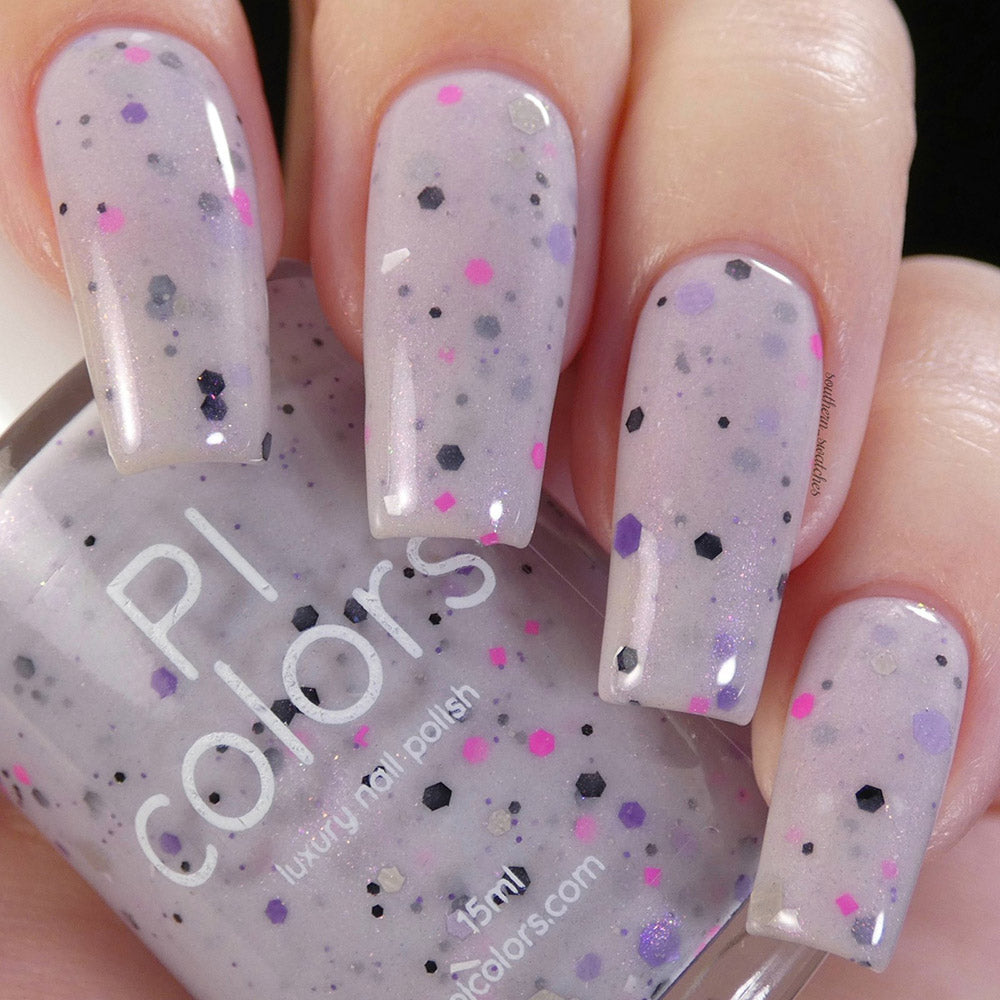 A close-up of manicured fingernails adorned with a speckled nail polish reveals a pale grayish purple crelly base enriched with small black and pink glitter flecks. The bottle of PI Colors' handmade Kirin.000 Lilac Purple Nail Polish is held, displaying the same captivating shade.