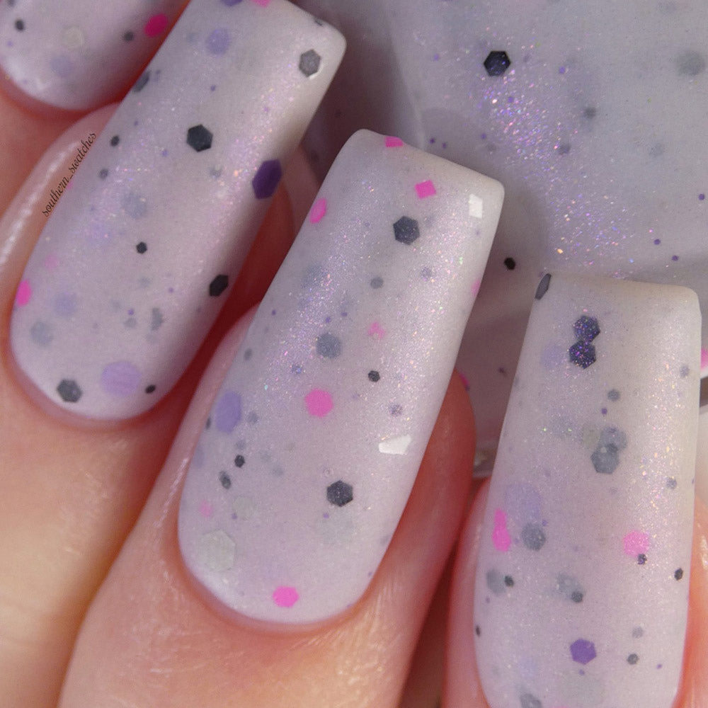 Here's a revised version of the sentence using the given product data:
A close-up of fingernails showcases PI Colors' handmade Kirin.000 Lilac Purple Nail Polish, featuring a long, glossy finish with a pale grayish purple crelly base and whimsical confetti-like speckles in black, pink, and purple. The polish also boasts a subtle shimmer.