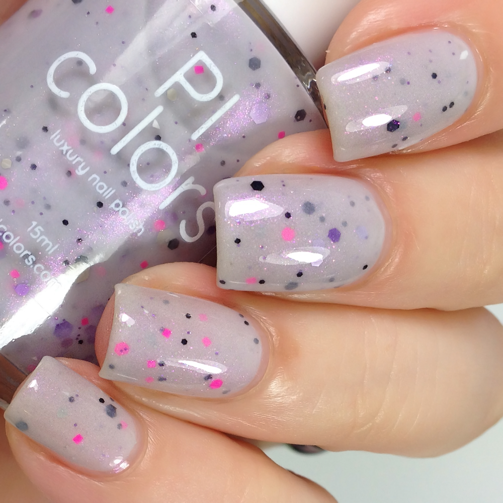 A close-up showcases a hand adorned with Kirin.000 Lilac Purple Nail Polish from PI Colors, which features black and pink speckles. This handmade polish has a hint of pale grayish purple crelly housed in a 15ml bottle, held gracefully between the fingers. The nails boast a glossy finish that enhances the shimmer effect.