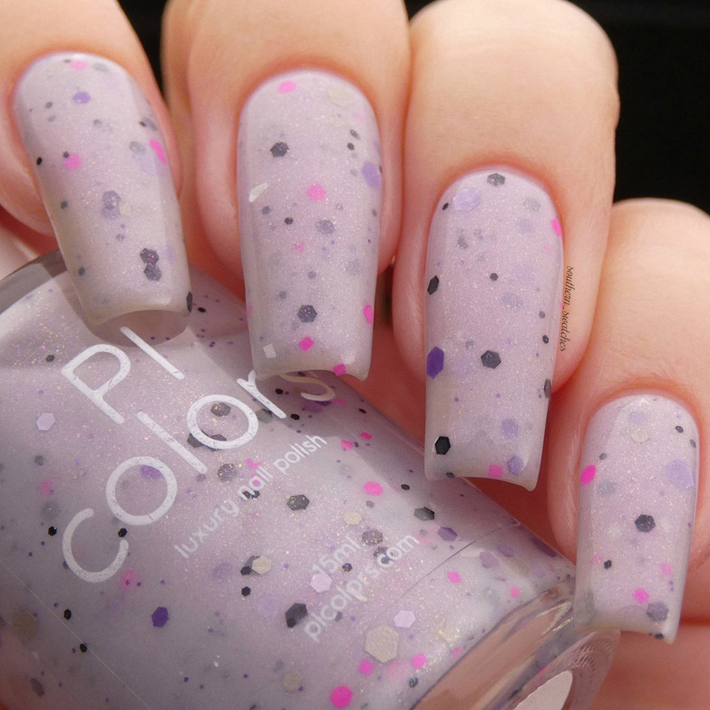 A close-up of a hand with custom batches of long nails painted in Kirin.000 Lilac Purple Nail Polish, featuring multicolored speckles in pink, black, gray, and purple. A bottle of PI Colors nail polish from the Animation Polish Pickup collection is held in the same hand.