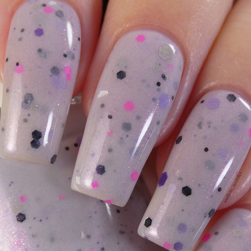 A close-up of fingers adorned with PI Colors' Kirin.000 Lilac Purple Nail Polish, a pale grayish purple crelly featuring black, pink, and white speckles in various hexagonal shapes. The finish is shiny with a subtle shimmer effect, highlighting the allure of this handmade polish.