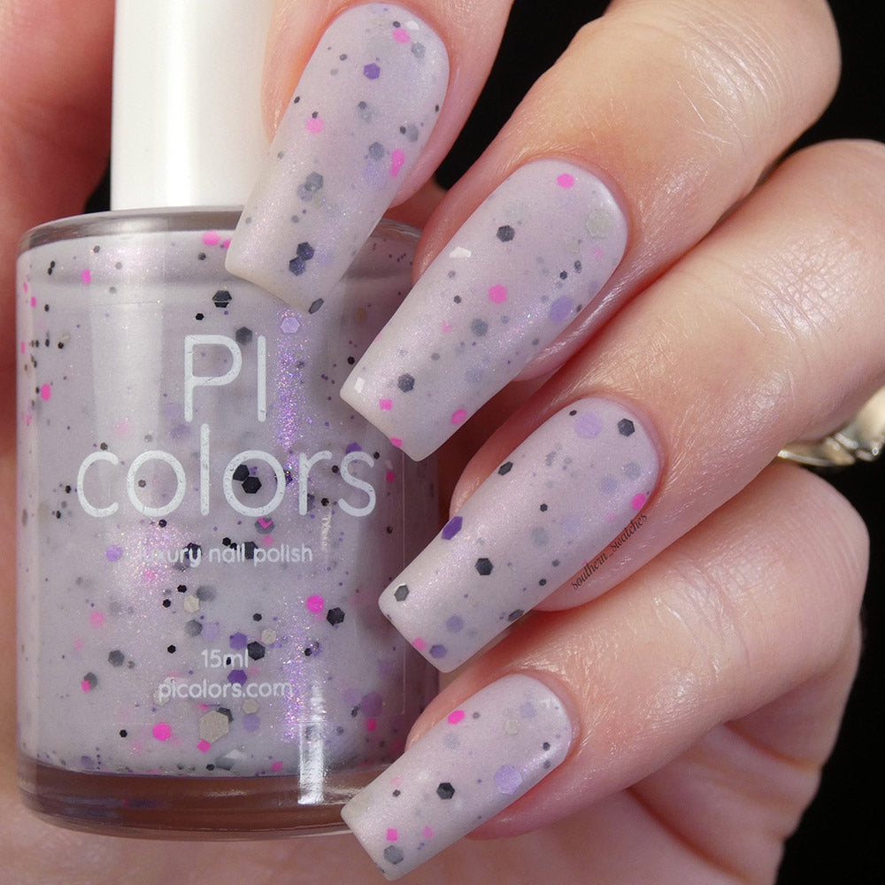 A hand with long square nails is painted in a pale grayish purple crelly, embellished with black, pink, and purple speckles. The bottle of the same handmade nail polish, labeled "Kirin.000 Lilac Purple Nail Polish" by PI Colors, is held between the fingers.