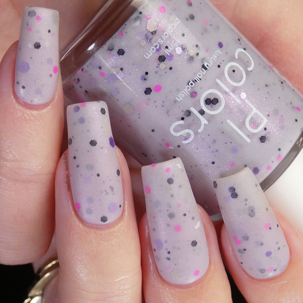 A close-up of a hand with long nails adorned in Kirin.000 Lilac Purple, a pale grayish purple crelly from PI Colors that features black, white, and pink speckles. The person holds a matching handmade nail polish bottle labeled "PI Colors.