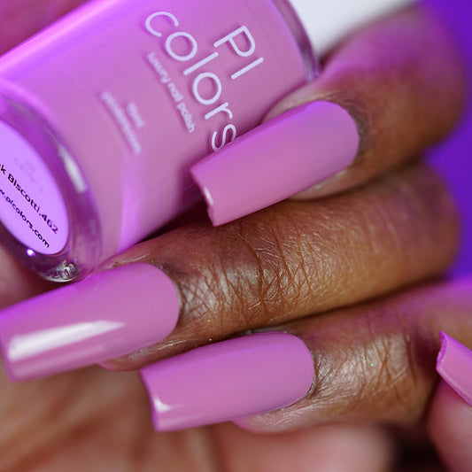 A hand with long, square-shaped nails painted in glossy Pink Biscotti.462 by PI Colors holds a matching handmade polish bottle. The background is blurred in purple tones.