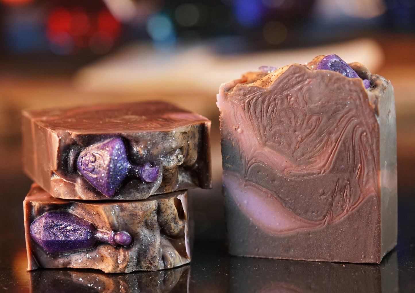 Three Potion Drip Artisan Soap Bars by PI Colors are displayed with swirling patterns and purple gemstone-like decorations. Two bars are stacked, one stands upright, beautifully showcasing their rustic texture and artistic design, inviting a luxurious bath experience on the reflective surface.