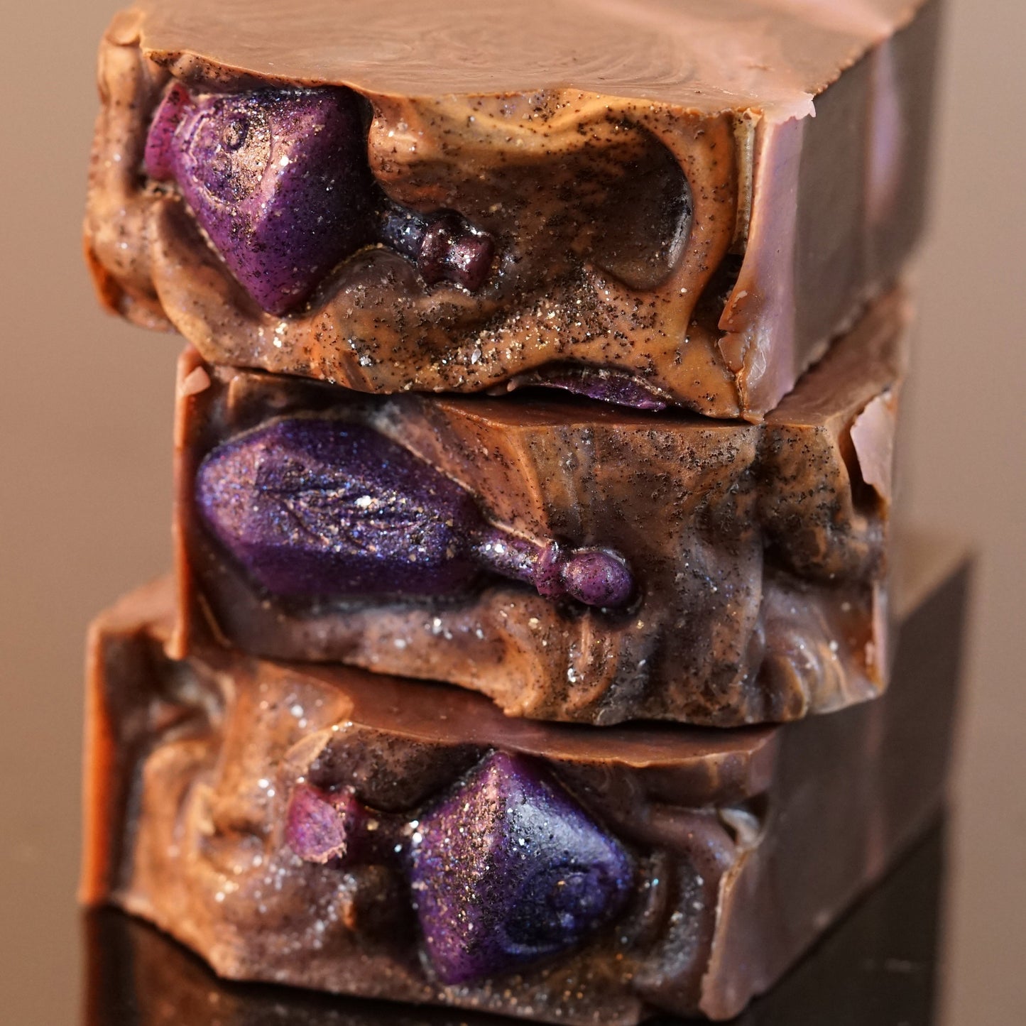 Three bars of PI Colors' Potion Drip Artisan Soap are stacked, each showcasing a glittery purple potion bottle design. The soap features swirled brown and purple hues and an earth-textured surface, promising a scented bath experience.