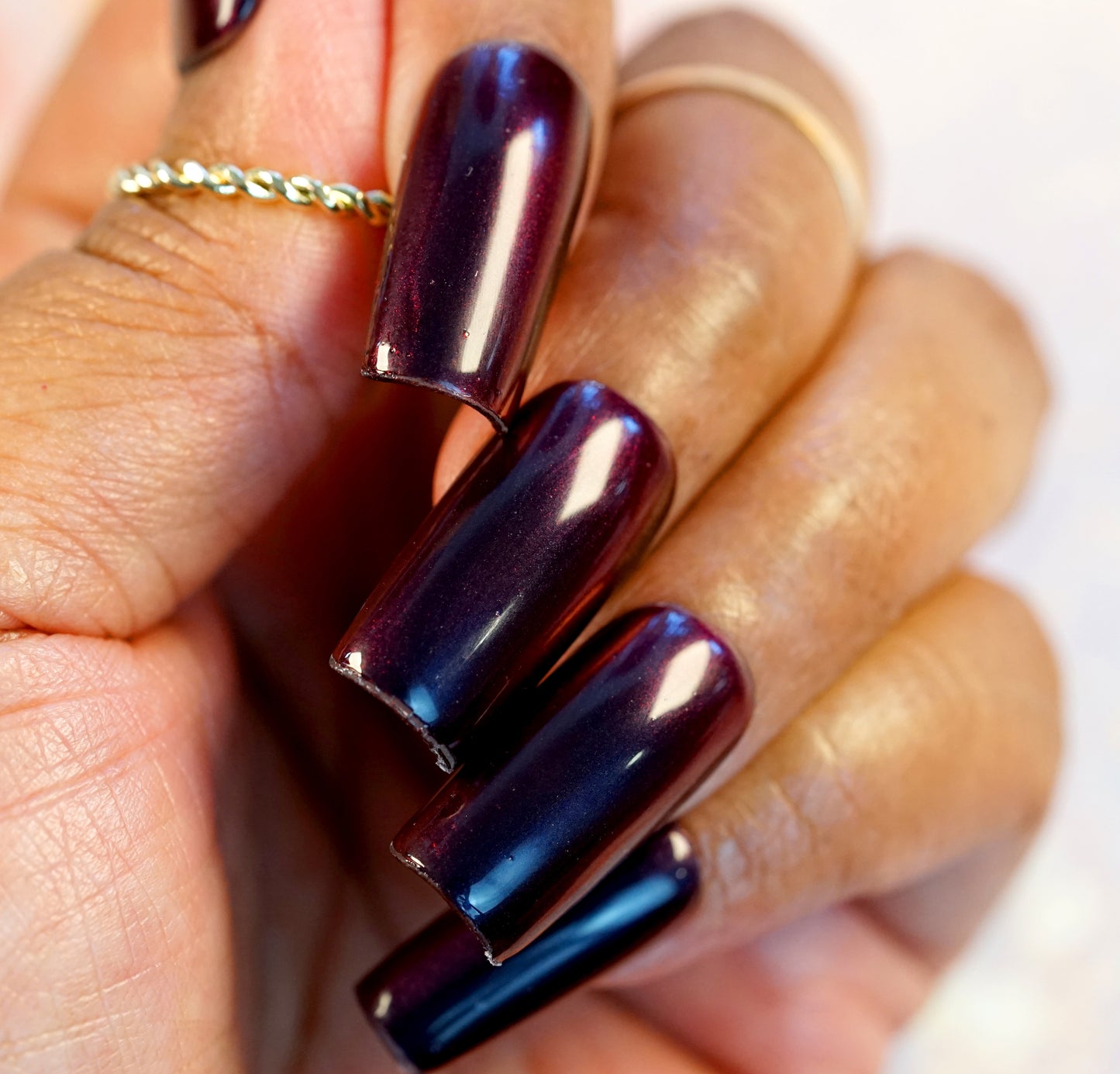 A close-up of a hand with long, glossy nails painted with PI Colors' Red Glow.295 nail polish, adorned with delicate gold rings. The luxurious lacquer shimmers in the light, revealing an intriguing depth to its shiny finish.