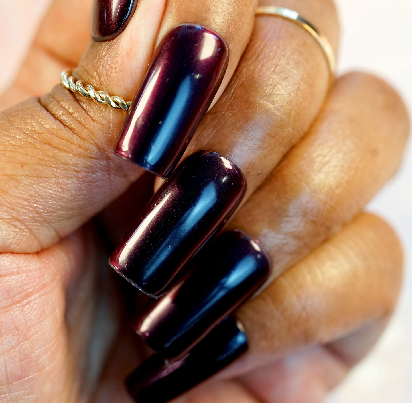 A close-up of a hand highlights long, sleek nails painted with PI Colors' Red Glow.295 Red Nail Polish in a rich burgundy hue. The medium brown skin tone beautifully complements the twisted gold ring on one finger. This 10-free formula guarantees the glossy finish is both stunning and safe.