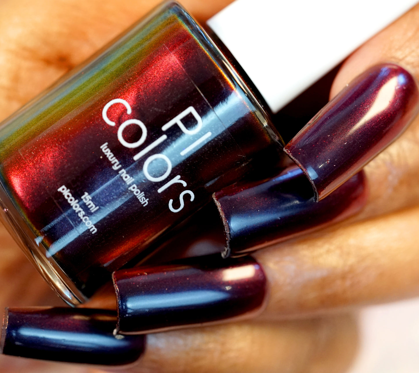 A hand with long nails painted in a luxurious, shimmering shade of "Red Glow.295" nail polish by PI Colors holds a matching bottle. The glossy finish catches the light, accentuating its deep, rich hue.