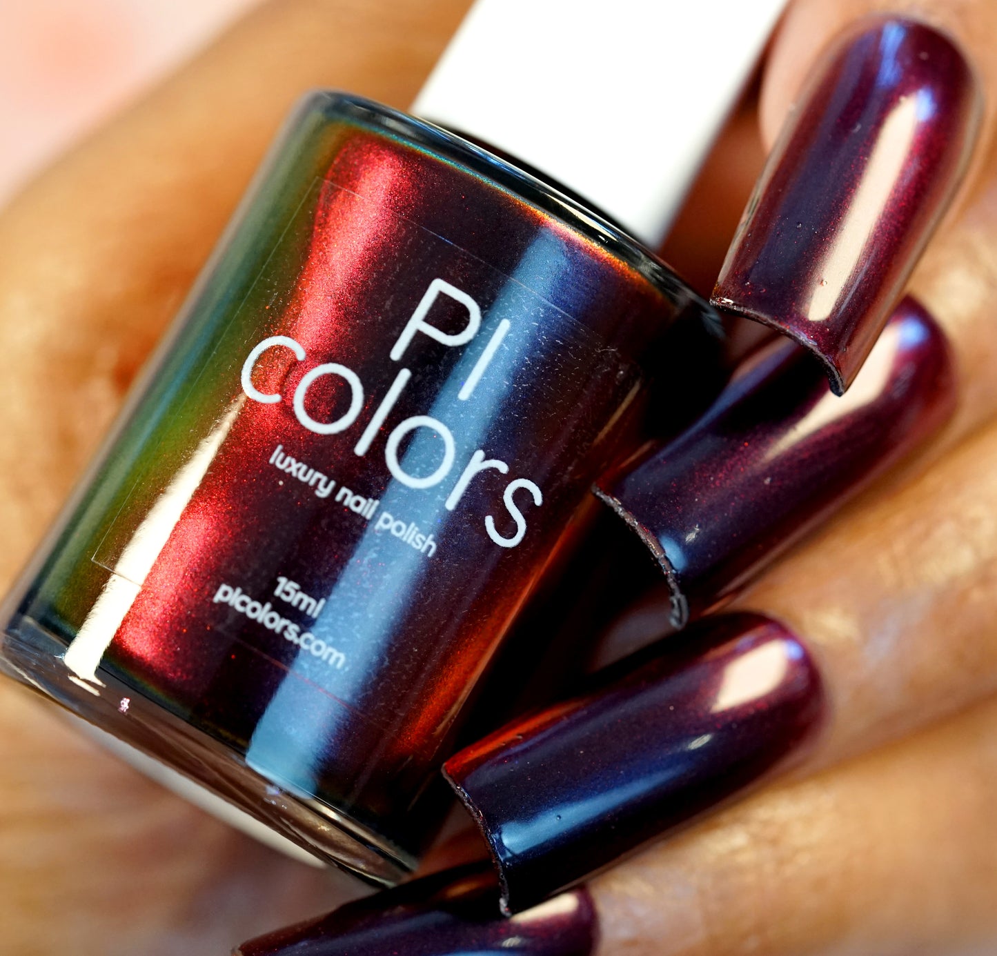 A close-up of a hand with nails painted in luxurious, color-shifting Red Glow.295 nail lacquer showcases a shiny dark burgundy hue. The hand holds a bottle of PI Colors nail polish in the same shade, featuring an iridescent finish that reflects elegant hues of deep red and green.