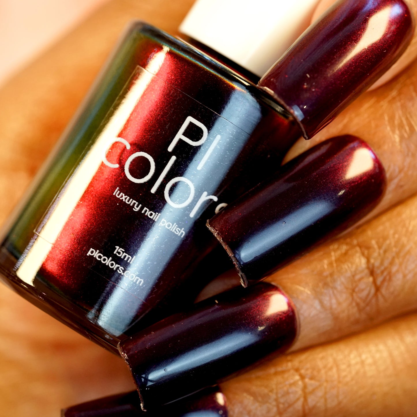 A close-up of a hand holding a bottle of PI Colors' Red Glow.295 nail polish in a glossy, dark burgundy shade. The polish on the nails matches the bottle color, showcasing a shiny finish that rivals any red nail polish.
