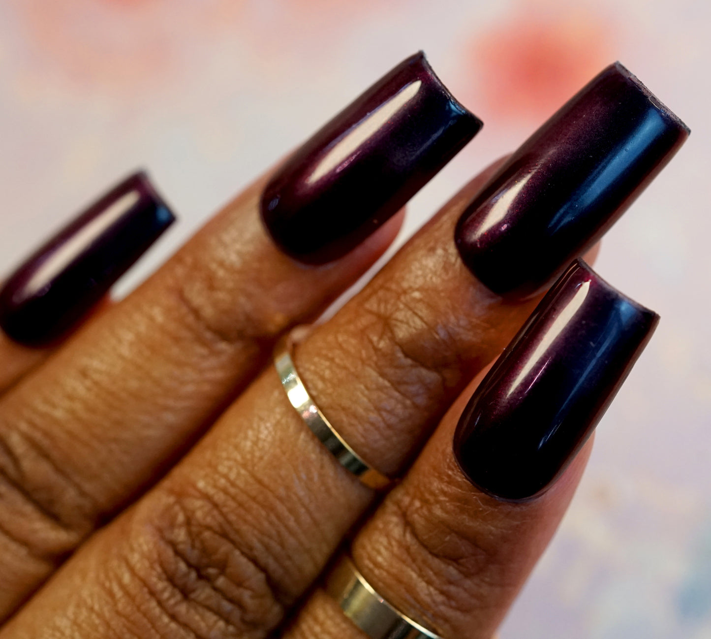 A close-up of a hand with long, dark purple glossy nails showcases stunning colorshift effects. The fingers, adorned with simple gold rings, capture the eye against a softly blurred background, highlighting the intricate detail and shine of the nails—all achieved with PI Colors' 10-free Red Glow.295 Red Nail Polish.