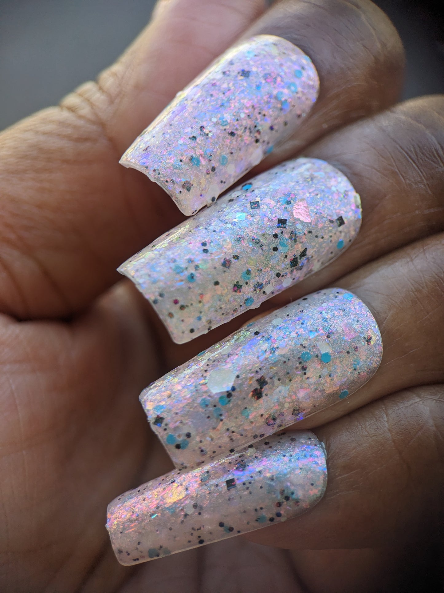 A close-up of a hand with long, square-shaped nails painted in PI Colors "Pink Ash.305" nail lacquer. The iridescent flakies create a dazzling, glossy finish, reflecting multicolored glitter in shades of blue, pink, and silver under the light for an eye-catching effect.