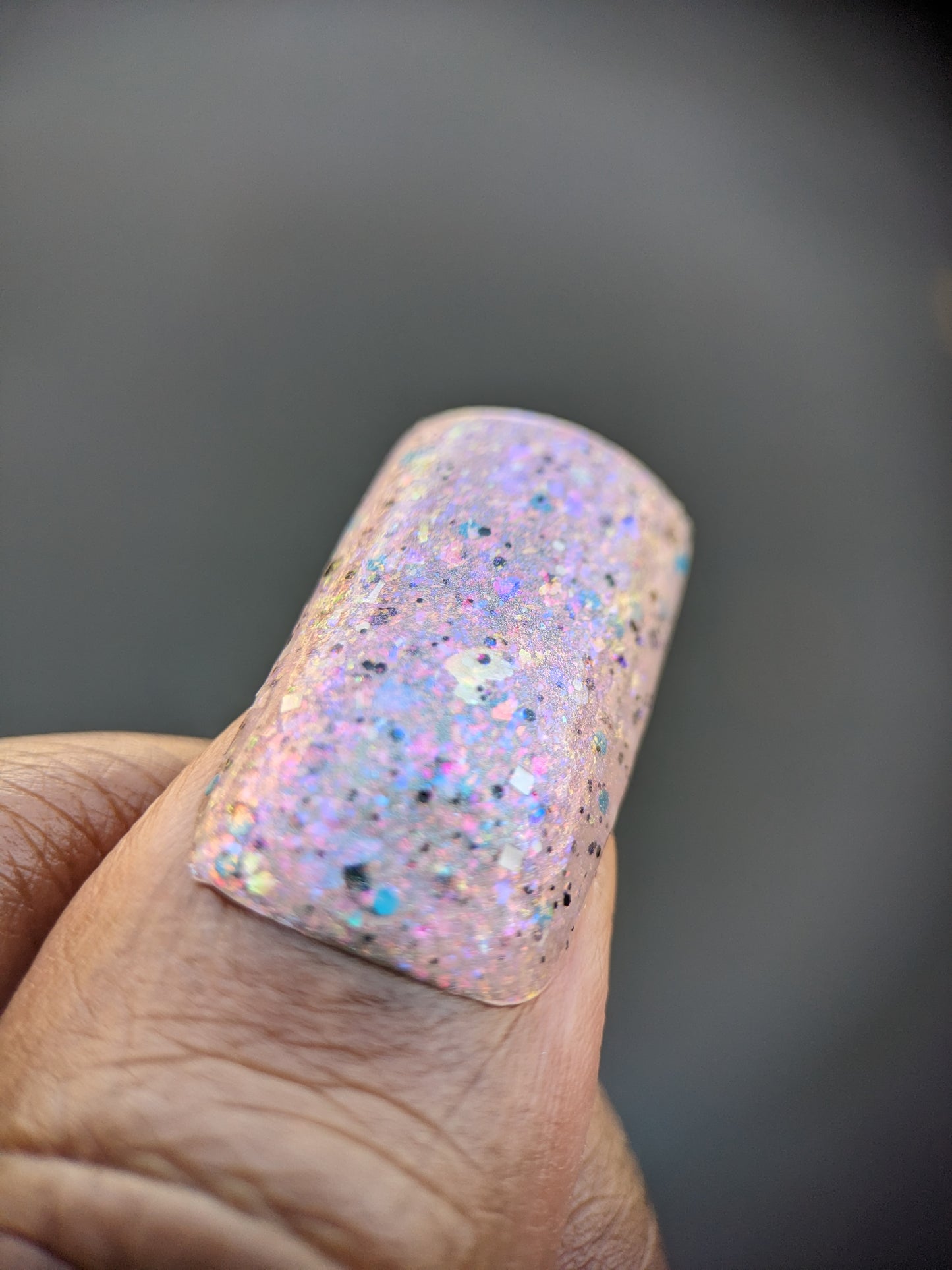 A close-up of a thumb adorned with PI Colors' Pink Ash.305 luxury nail lacquer in pastel hues. The iridescent flakies shimmer under light, displaying a blend of pink, purple, and blue. The angled thumb highlights the color's texture beautifully.