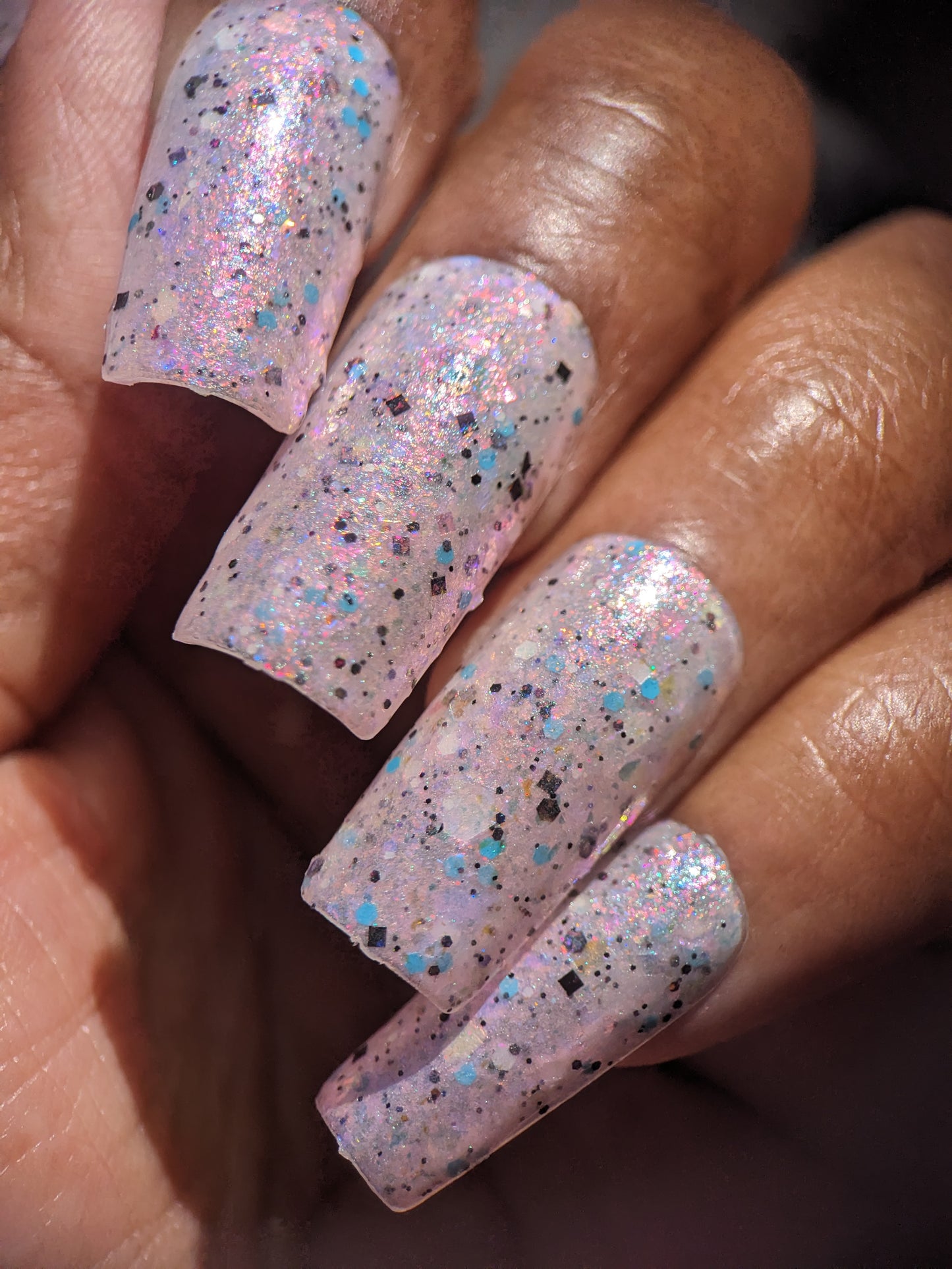 A hand with long fingernails painted in PI Colors' luxurious Pink Ash.305 nail lacquer shimmers with iridescent flakies and multicolored glitter. The pink and blue shades catch the light beautifully, complementing the rich brown skin tone.