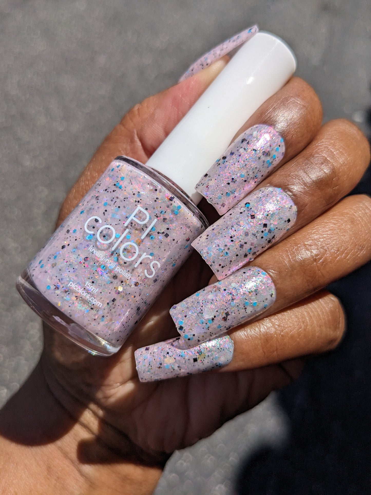 A hand with long nails displays PI Colors' Pink Ash.305, a luxury nail lacquer. The speckled pastel pink polish sparkles with iridescent flakies and multicolored glitter on a shimmery base, dazzling in the sunlight outdoors.