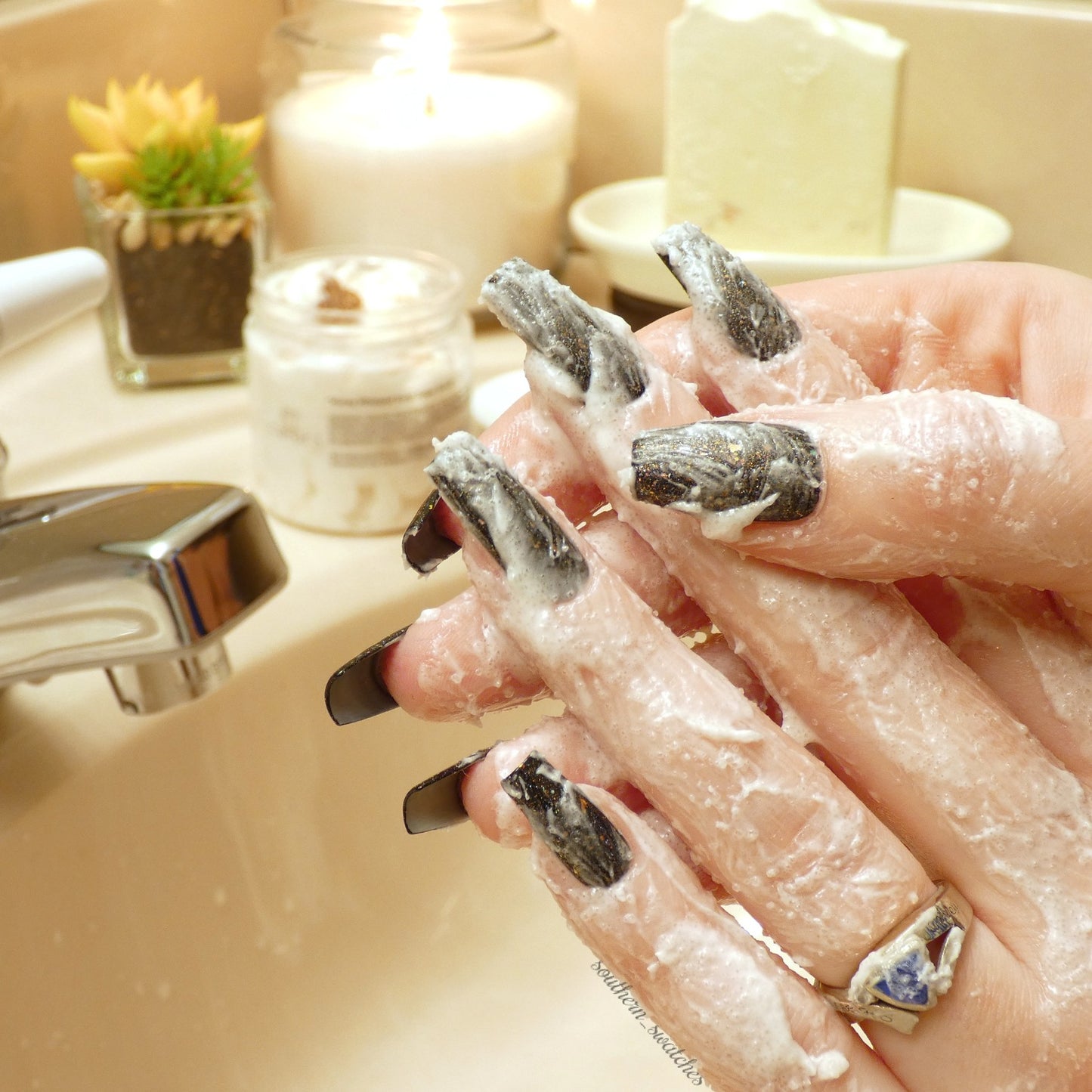 A close-up of hands with long, dark-painted nails being lathered with the creamy PI Colors Honey Scented Whipped Soap Sugar Scrub over the sink. A lit candle, potted plant, and skincare products enhance the soothing bathroom setting as luxurious bubbles offer gentle exfoliation in this tranquil moment.