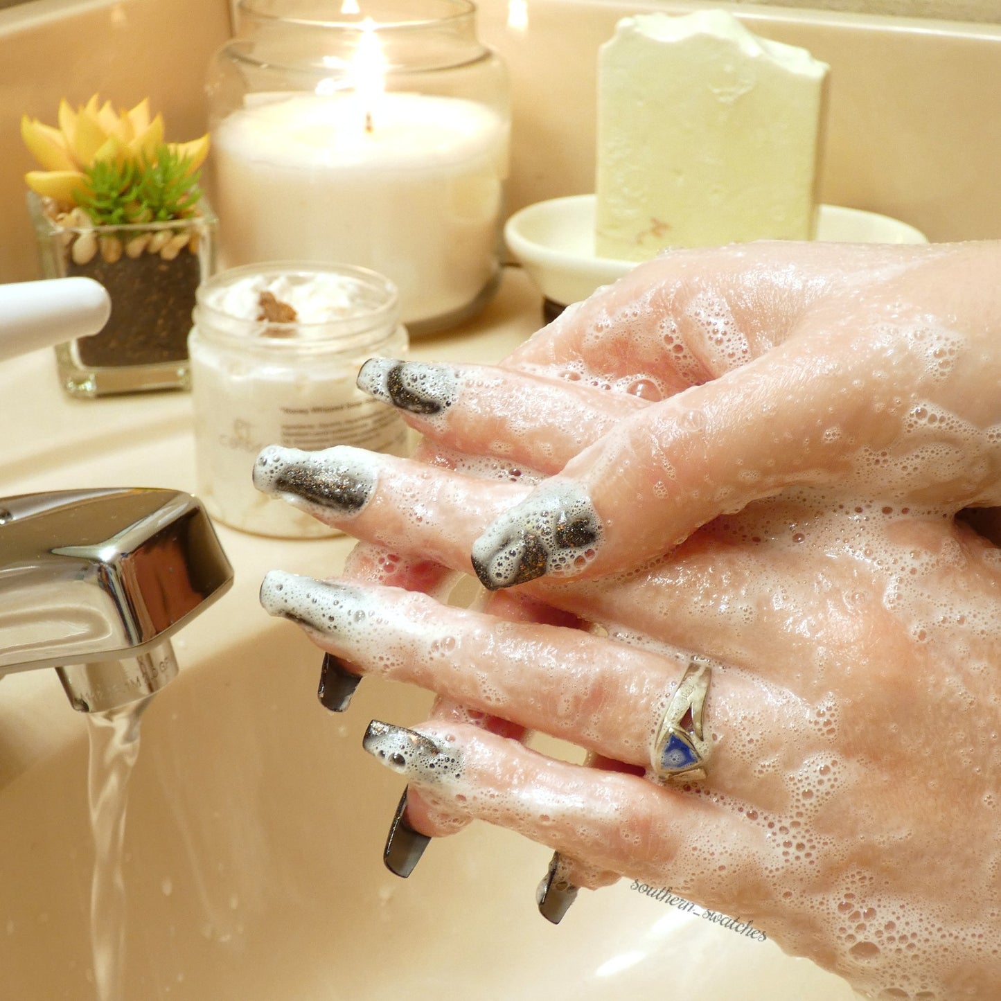 Glittery nail-polished hands are covered in PI Colors' Honey Scented Whipped Soap Sugar Scrub under running water at a sink, with a candle and a small plant gracing the countertop in the background.