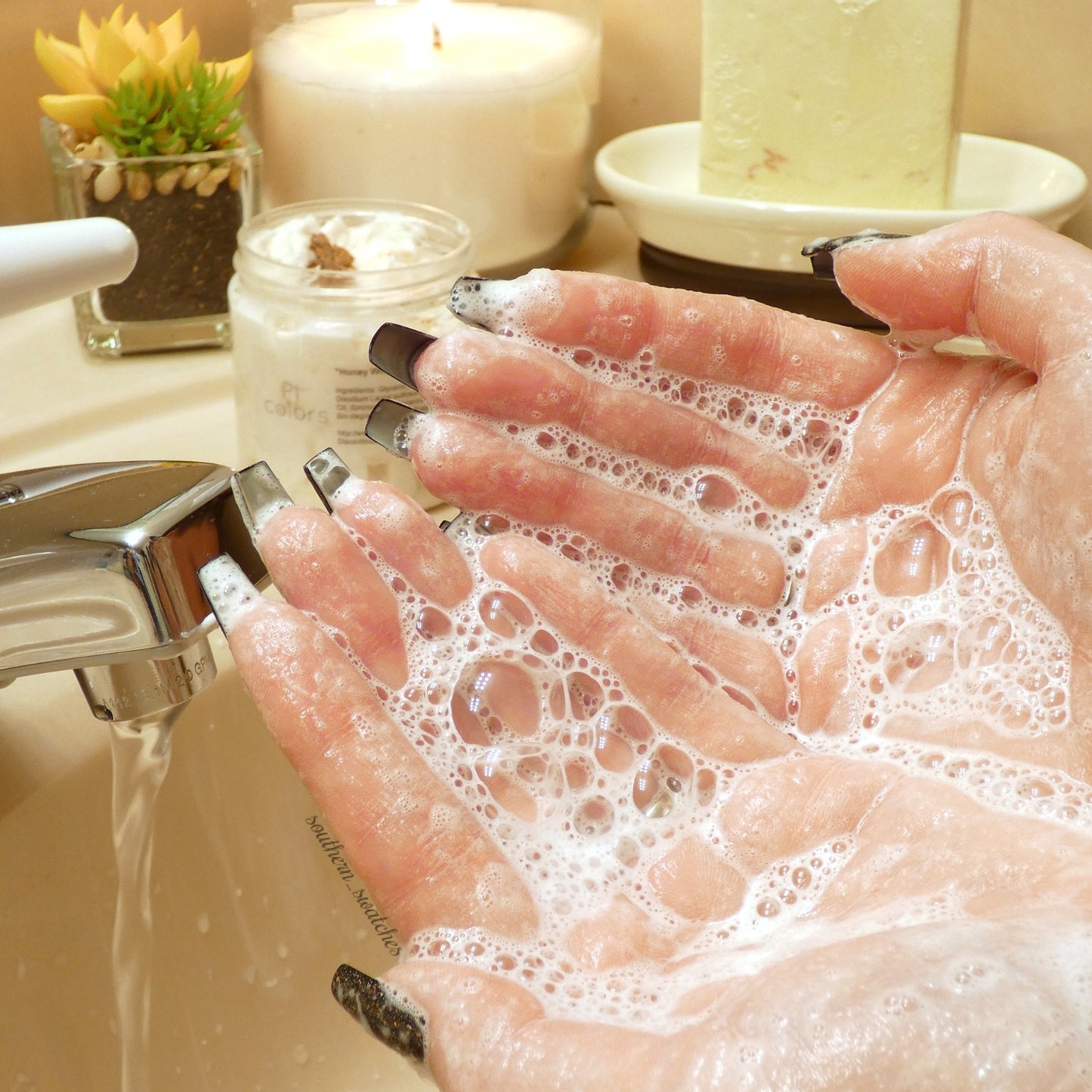Close-up of hands with black-painted nails lathered in PI Colors' Honey Scented Whipped Soap Sugar Scrub under running water from a bathroom faucet. In the background, a lit candle, succulent plant, and soap container create a serene setting.