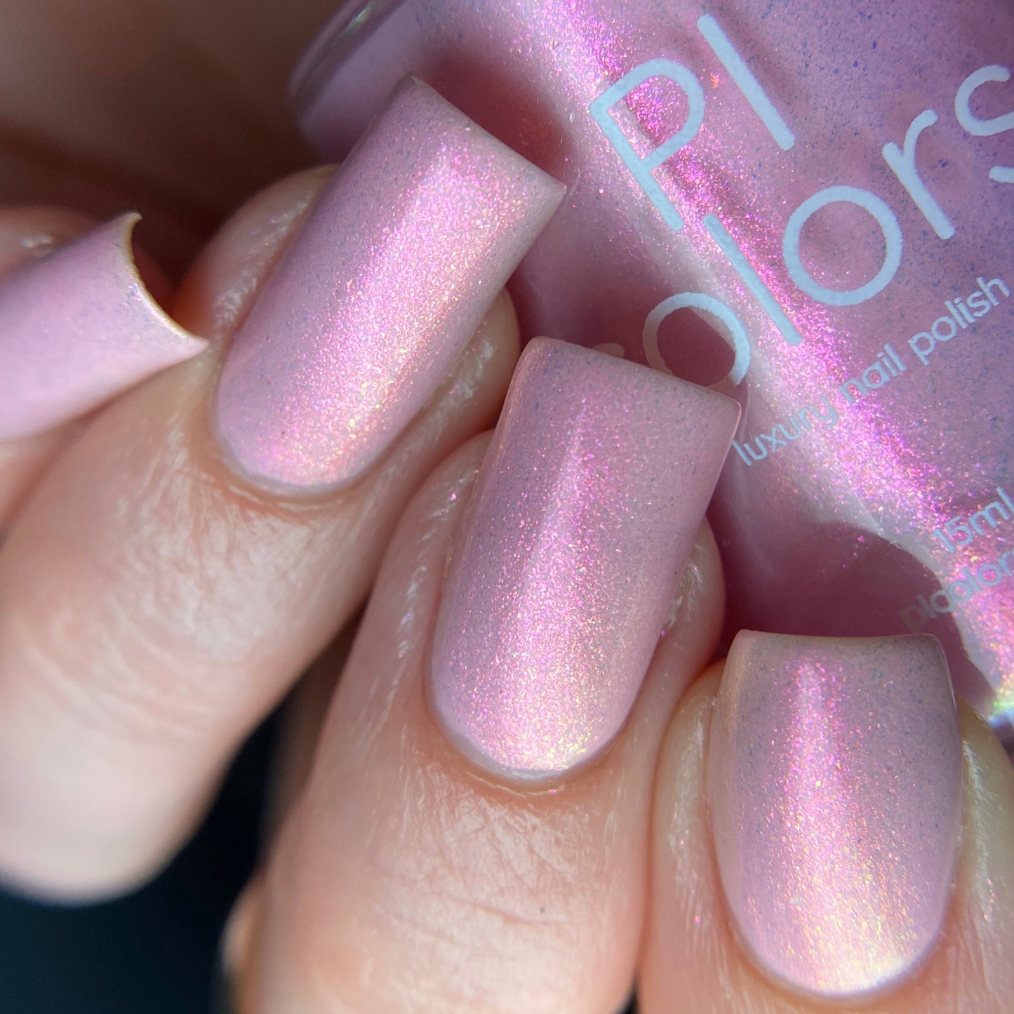A close-up captures a hand with elegant, long nails painted in Sunset Aurora.322, a luxurious pink shimmer nail polish by PI Colors. In the background, the handmade bottle perfectly complements the iridescent hue on the nails.