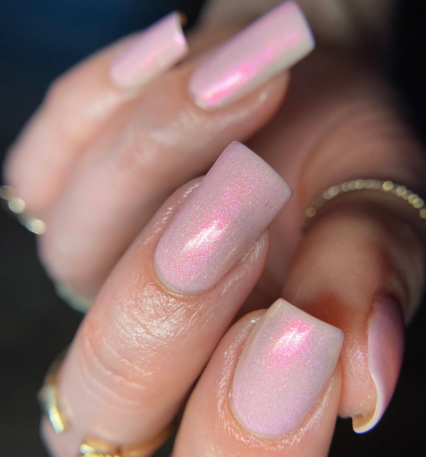 A close-up of a hand with long, square-shaped nails showcases the "Sunset Aurora.322 Pink Nail Polish" by PI Colors, shimmering in iridescent pale pink hues. The nails gleam as they catch the light, revealing subtle hints of lavender. Several gold rings add a touch of elegance to the fingers.