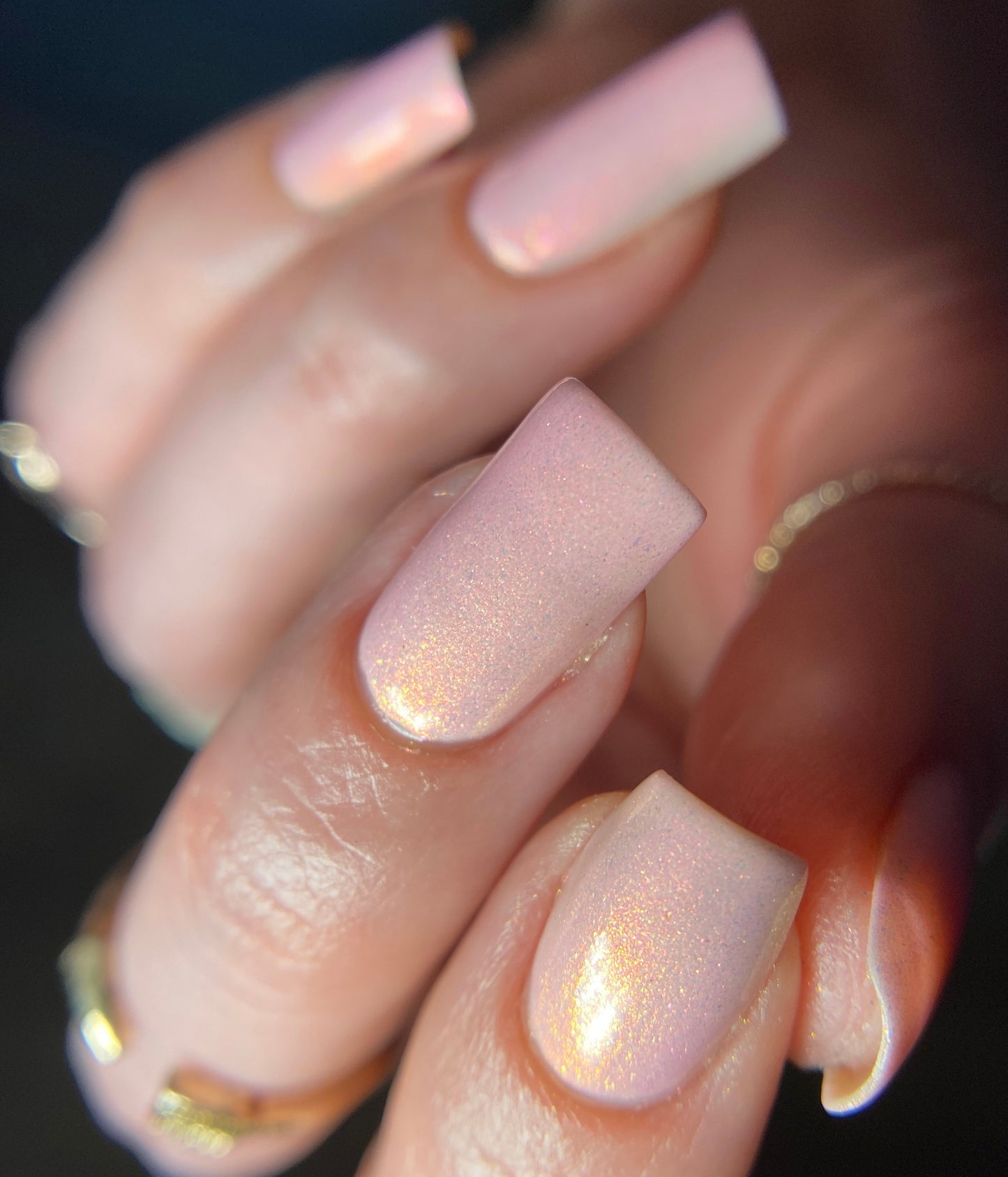 A close-up showcases a hand flaunting freshly manicured nails covered in PI Colors' Sunset Aurora.322, a glossy, pearlescent pink nail polish. The square-shaped nails subtly reflect light, complemented by gold rings, highlighting the luxurious quality of this 10-free luxury lacquer.