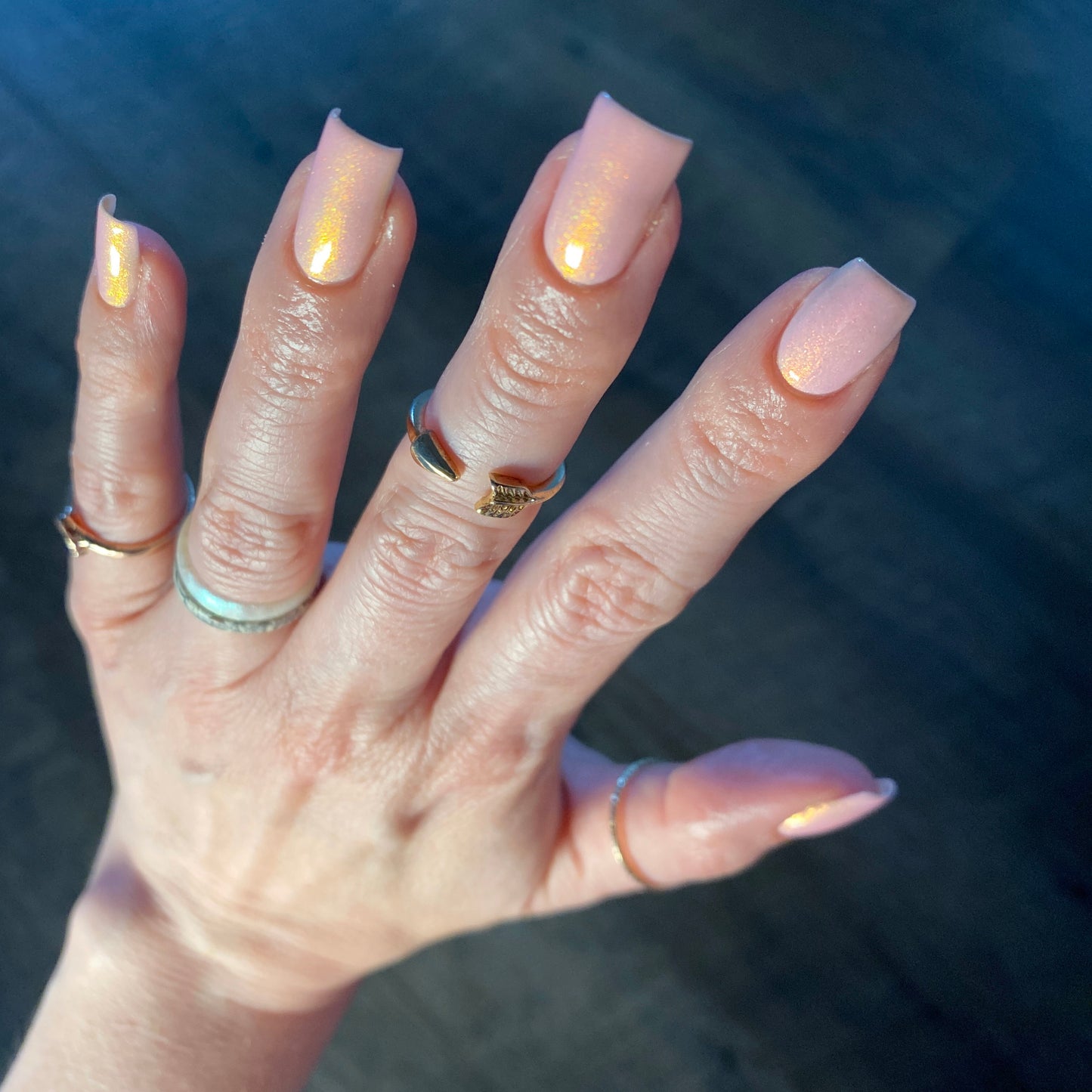 A hand flaunts long, glossy nails painted in PI Colors' Sunset Aurora.322 Pink Nail Polish, characterized by a peach hue with a pink shimmer. The subtle iridescent sheen enhances the luxurious appearance of the lacquer, while several thin gold and silver rings decorate the fingers against a dark, textured backdrop.