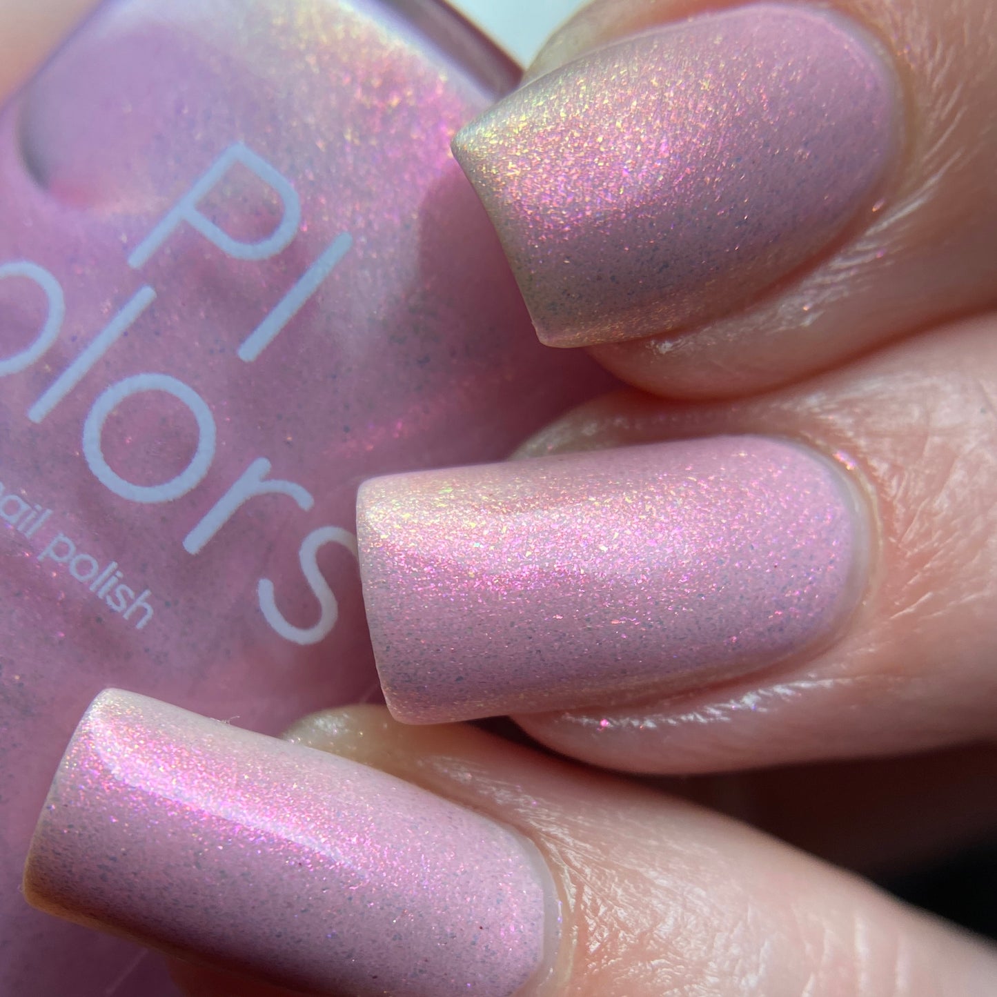 Close-up of manicured nails painted in PI Colors' Sunset Aurora.322 Pink Nail Polish. The glossy finish reflects light, showcasing a soft, iridescent sparkle. A bottle of this 10-free luxury lacquer is nearby, partially visible in the background.