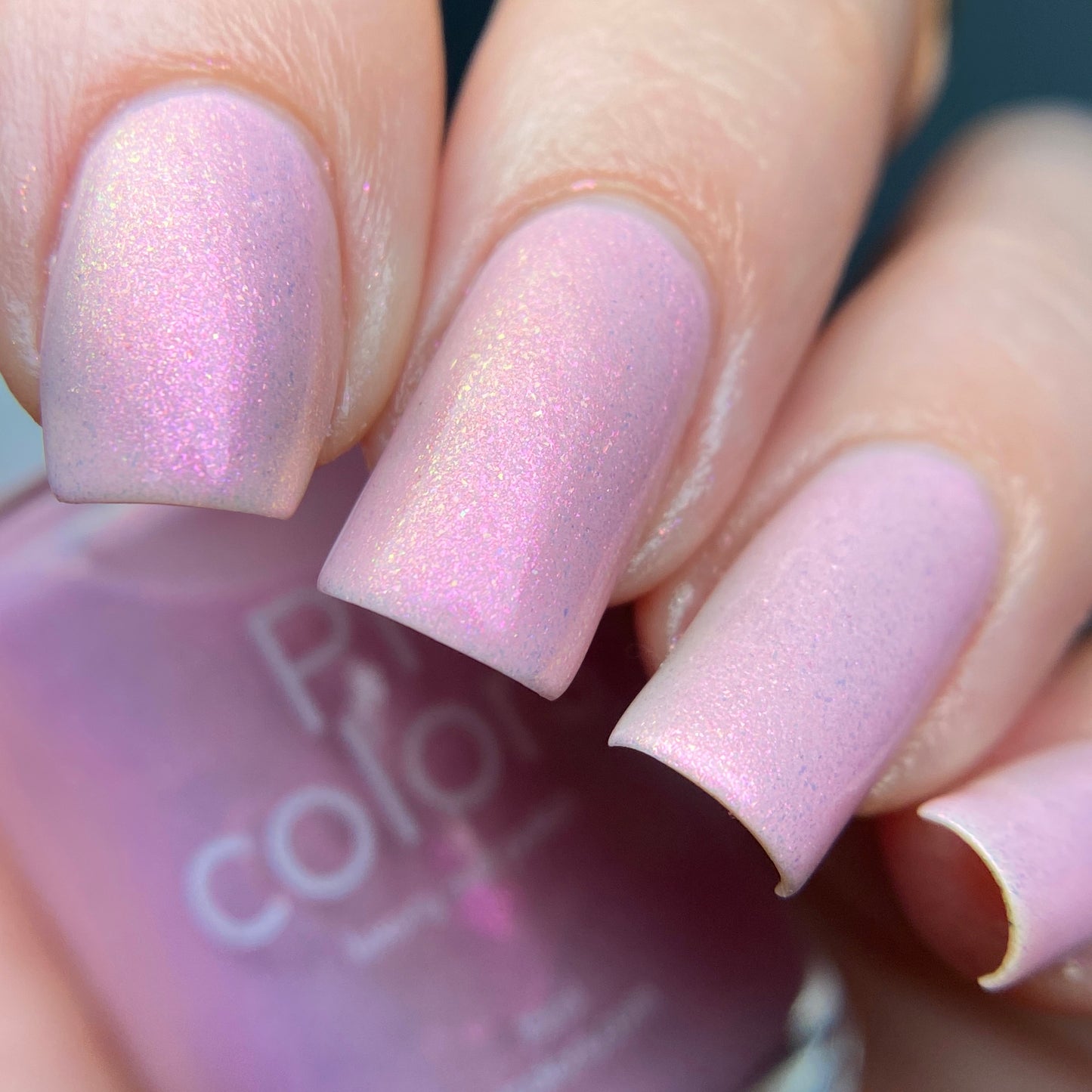 A close-up shows fingers adorned with square-shaped nails painted in the luxurious Sunset Aurora.322 Pink Nail Polish by PI Colors. The shimmering pastel pink reflects light beautifully, highlighting its glossy finish and subtle sparkle, while the blurred background accentuates the elegance of this 10-free luxury lacquer.