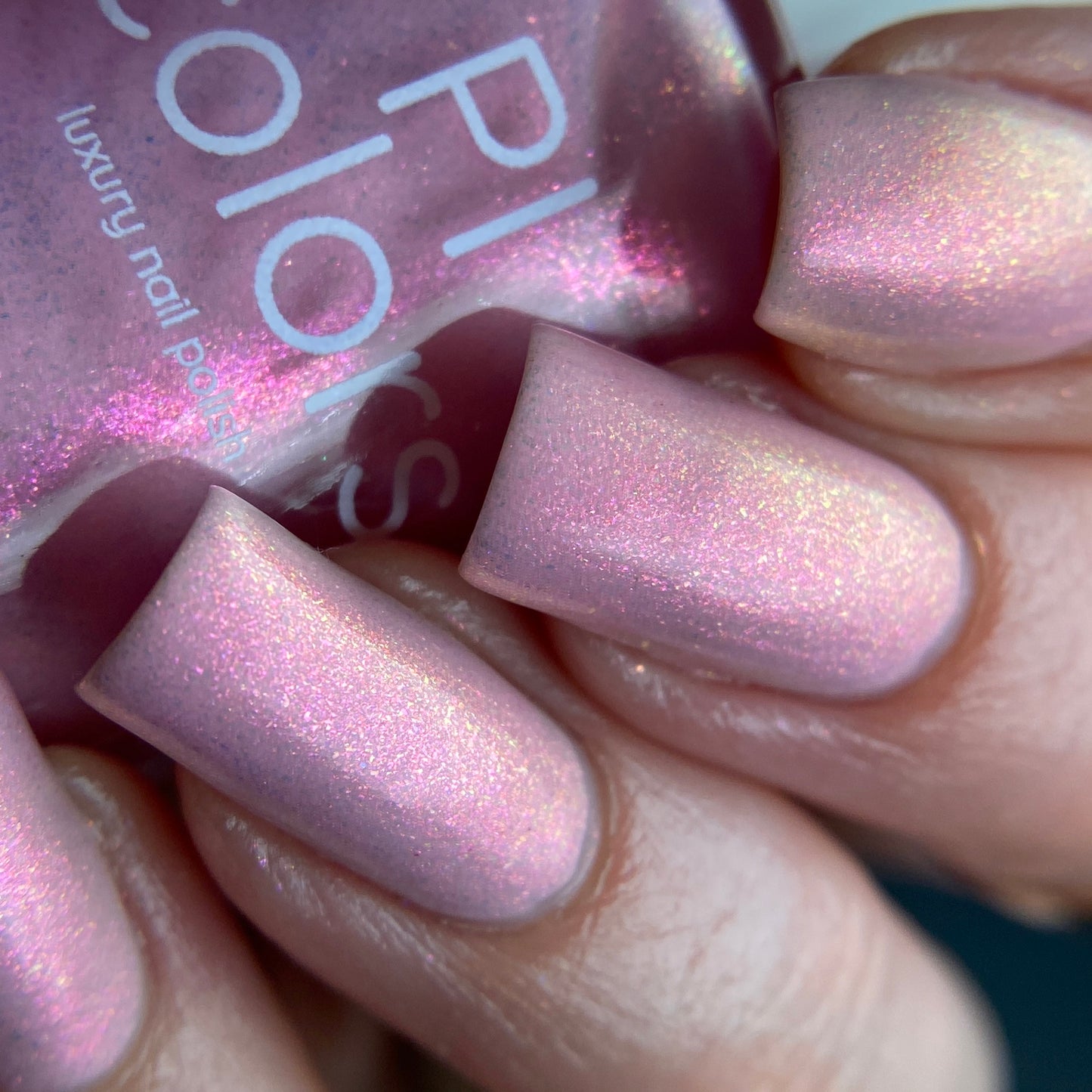 A close-up of a hand flaunts long, square-shaped nails adorned with PI Colors' Sunset Aurora.322 Pink Nail Polish in an iridescent pink shimmer. In the background, a partially visible bottle showcases this same mesmerizing shade, enhancing the elegant allure of this handmade nail polish masterpiece.
