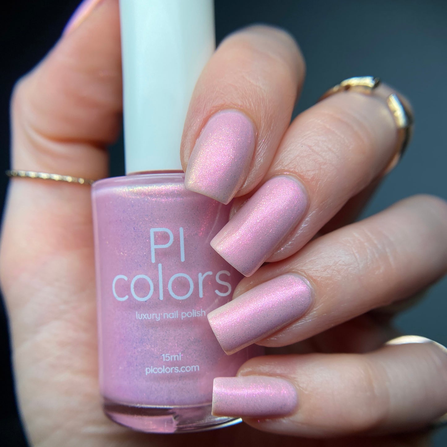 A hand adorned with rings holds a bottle of PI Colors Sunset Aurora.322 Pink Nail Polish, a 10-free luxury lacquer. The nails, painted in a shimmering pink that perfectly matches the handmade polish in the bottle, are beautifully highlighted against the softly blurred background.
