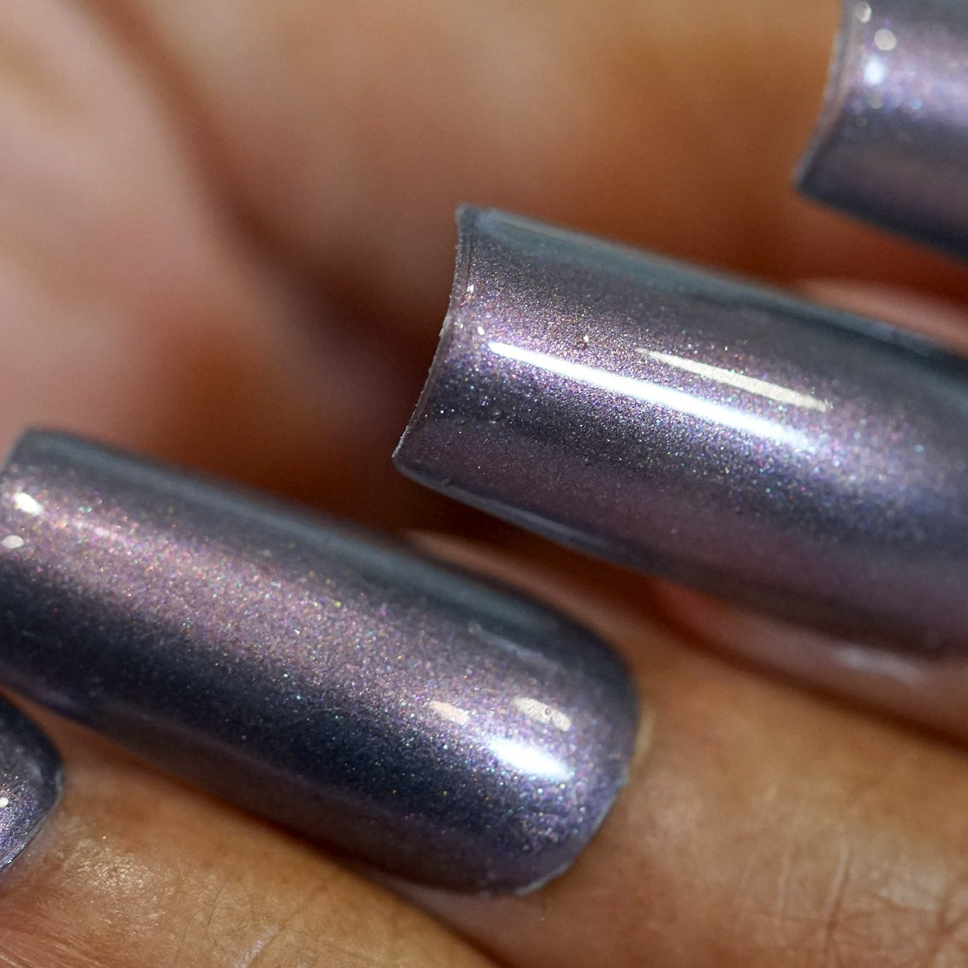 A close-up of a hand with 10-free, Twilight Moon.068 nails from PI Colors flaunts a metallic purple hue with a glossy finish and subtle shimmer. The long, well-manicured nails accentuate the reflective, iridescent quality of the muted gray purple polish.