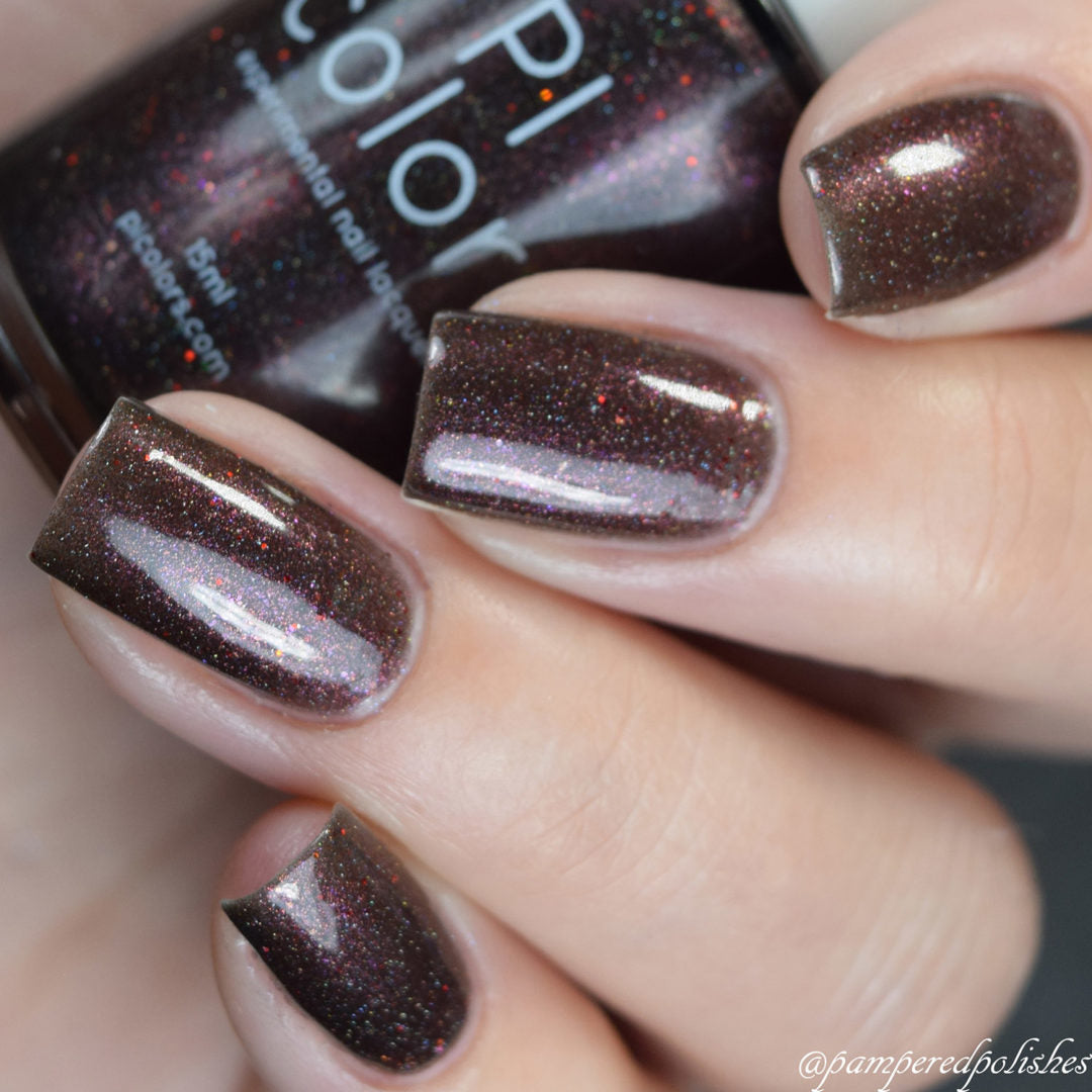A hand with short, neatly painted nails in Ursidae.072 Red Black Nail Polish from PI Colors is enhanced by holographic powders that shimmer with fine glitter. The 10-free polish bottle, partially visible, perfectly matches the nail color. The skin tone is light.