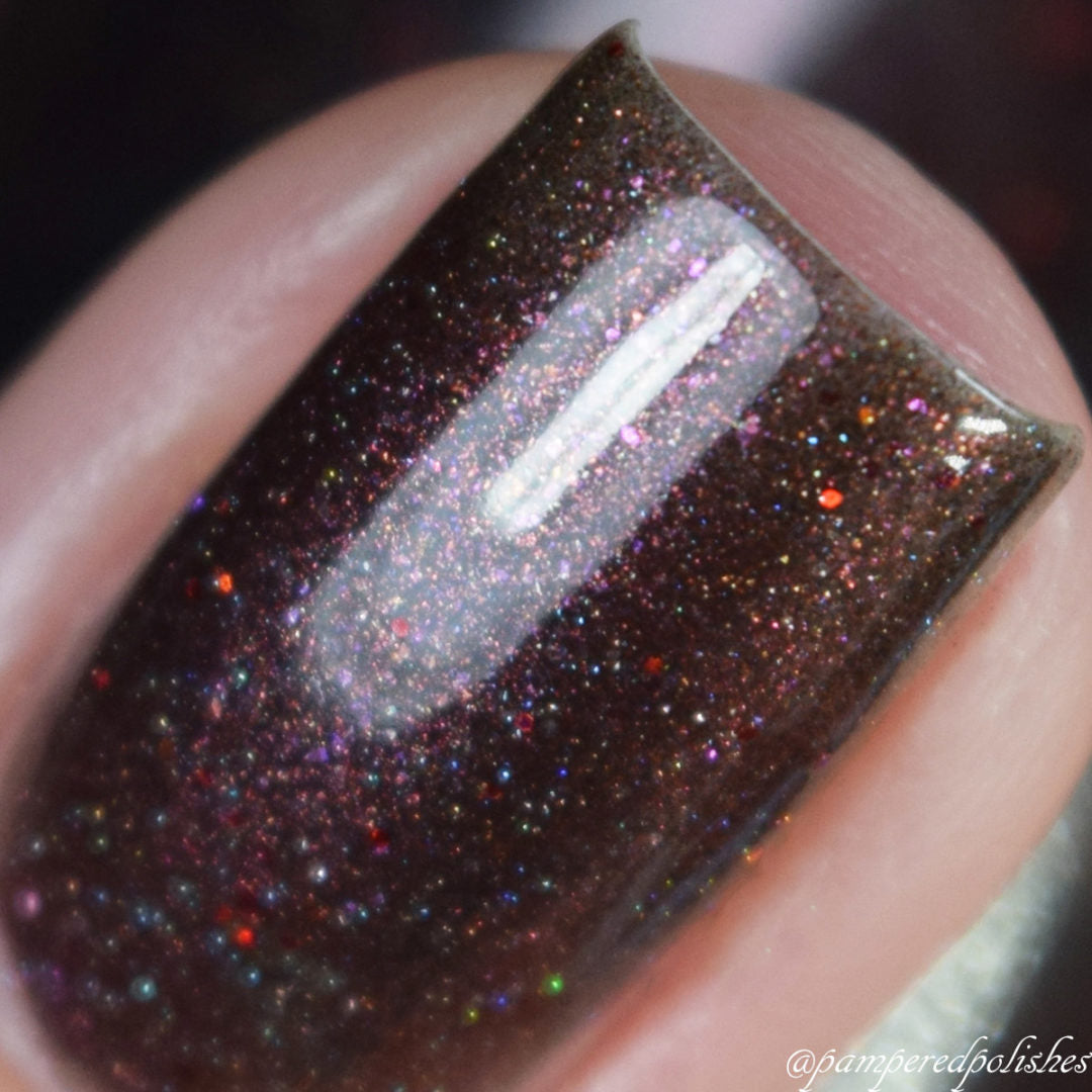 Close-up of a fingernail adorned with Ursidae.072 Red Black Nail Polish by PI Colors, showcasing dark brown polish with multicolored, shimmering glitter particles and holographic powders. The glossy finish reflects light, accentuating the vibrant red and pink specks.