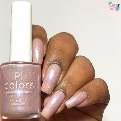 A hand with long nails painted in the shimmery, pale pink Moazagotl.127 Pink Nail Polish holds a bottle of the same polish. The bottle features "PI Colors" in white text, and the glittery holographic glitters give it an iridescent finish.