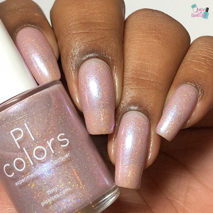 A hand with long nails painted in shimmery, iridescent "Moazagotl.127 Pink Nail Polish" is displayed. The handmade bottle from PI Colors is held in the other hand. This 10-free nail polish features holographic glitters for a glittery finish and a soft, ethereal look.