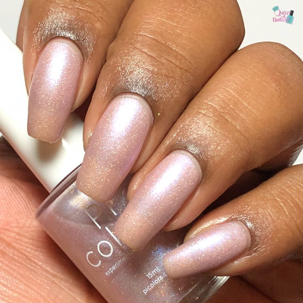 A close-up of a hand with long nails painted in Moazagotl.127 Pink Nail Polish from PI Colors showcases the elegance of 10-free nail polish. The matching bottle is gracefully held, partially visible, while the medium to dark skin tone offers a striking contrast to the light hues adorned with delicate holographic glitters.