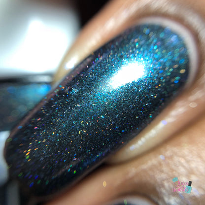 Close-up of a polished fingernail showcasing Noctilucent.111 Black Nail Polish by PI Colors, featuring a shimmering dark blue hue with holographic powders. The polish beautifully reflects light, creating a vibrant, sparkling effect. The blurred background accentuates the intricate nail detail.