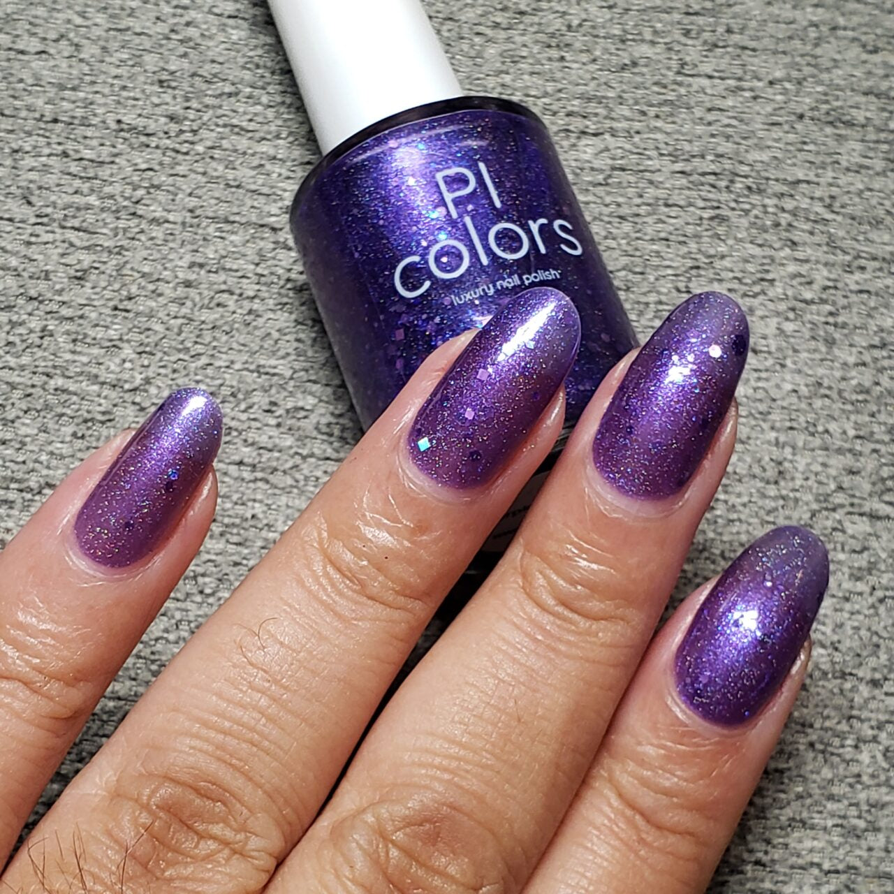 A hand with long nails painted in a dazzling holo purple glitter rests on a gray textured surface. Nearby, a bottle of PI Colors' Purple Smoke.001 Purple Nail Polish showcases the same sparkling shade as the bold purple jelly polish.