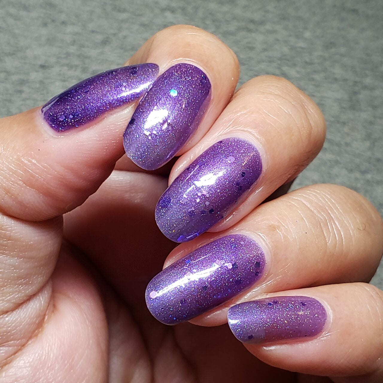 Close-up of a hand showcasing long, almond-shaped nails embellished with Purple Smoke.001 Purple Nail Polish by PI Colors, featuring a glossy jelly finish with holographic purple glitters. The textured gray background enhances the elegance of the Luxury Nail Lacquers.