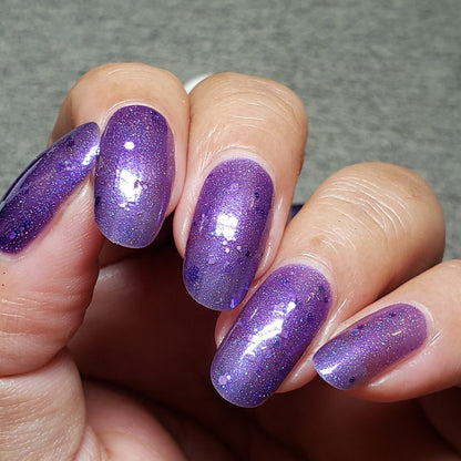 A hand exhibits nails adorned with PI Colors' Luxury Nail Lacquer called Purple Smoke.001, an iridescent purple jelly polish. The glossy finish features tiny holo purple glitters, with a neutral gray background emphasizing the vibrant nail color.