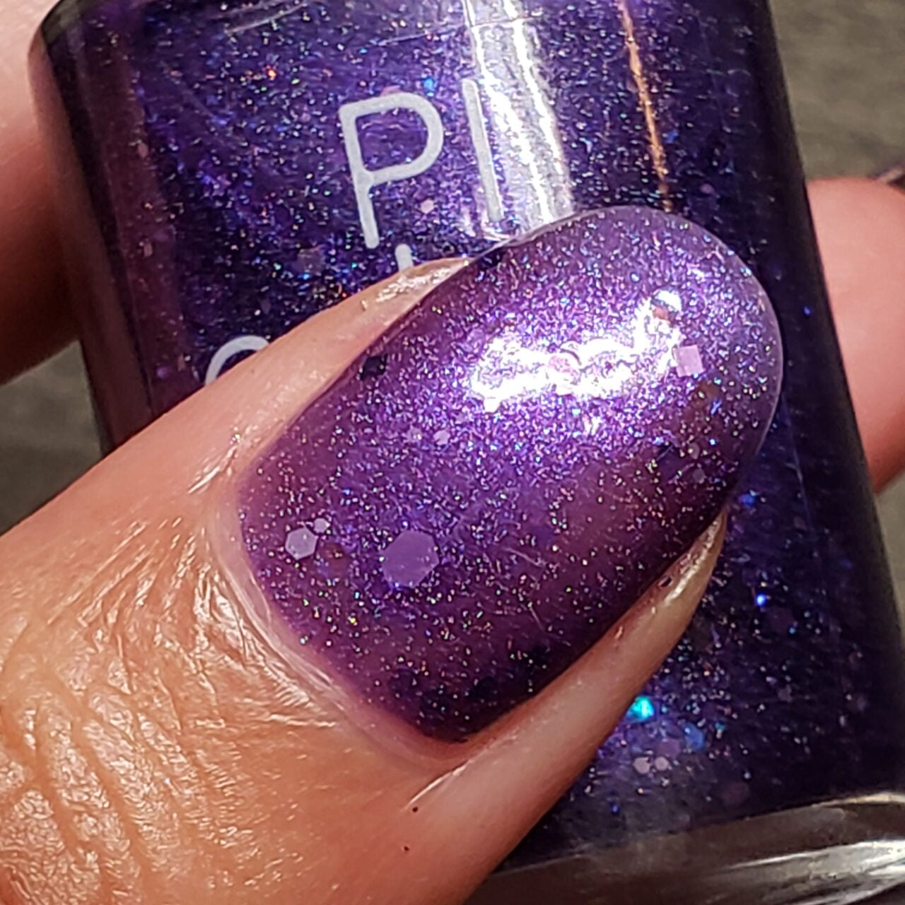 A close-up shot of a fingernail coated in shimmery purple jelly polish with holographic purple glitters, as the person holds a bottle of PI Colors' Purple Smoke.001 Purple Nail Polish, with part of the brand name subtly visible.