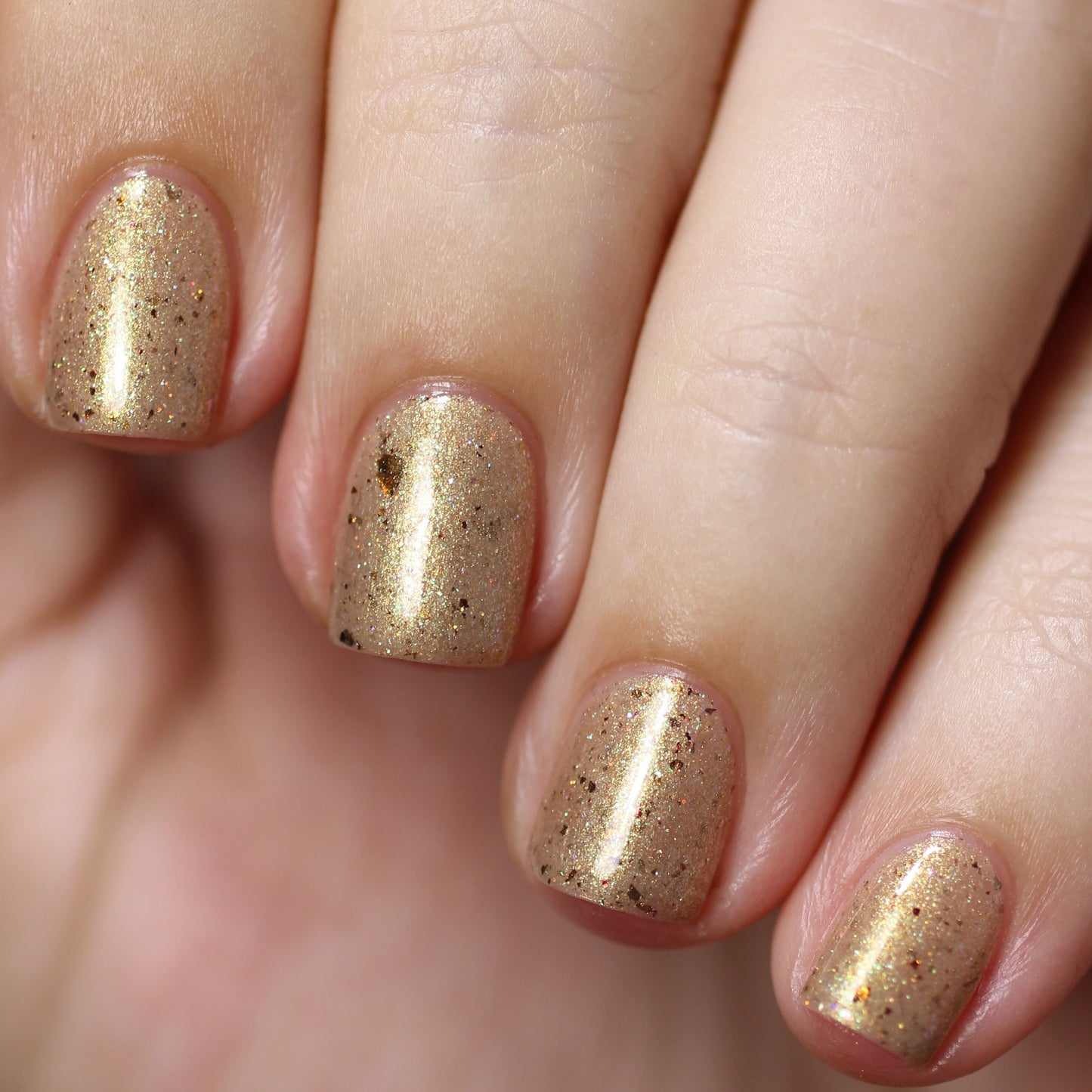 A close-up of a hand with short nails painted in PI Colors' Golden King.001 Gold Nail Polish showcases a luxurious shimmering gold with flecks of black. Enhanced by a glossy finish and holographic glitter, the skin appears smooth and well-groomed.