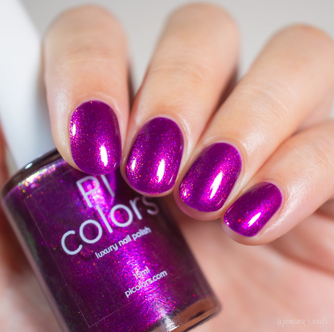 A hand with short, neatly manicured nails painted in shimmering, vibrant deep purple holds a matching bottle of PI Colors Aubergine.100 luxury nail lacquer.