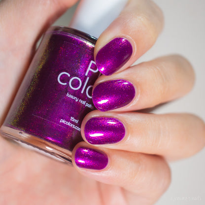 A hand with neatly manicured nails painted in a sparkly, metallic magenta shade is holding a bottle of PI Colors luxury nail lacquer labeled "Aubergine.100.