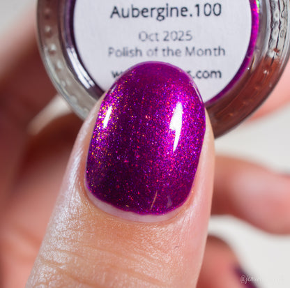 A close-up of a fingernail painted with vibrant, shimmery deep purple PI Colors Aubergine.100 nail polish. In the background is a bottle labeled "Aubergine.100, Polish of the Month" from PI Colors, highlighting this luxury lacquer.