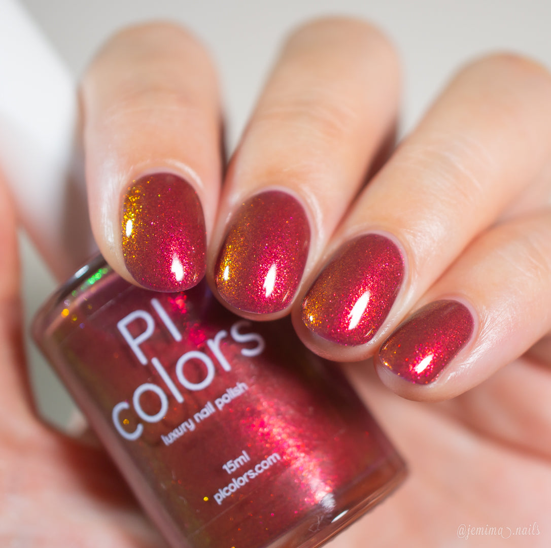 A hand with neatly manicured nails painted in Autumnal Equinox.100, a shimmering red and gold multichrome by PI Colors, holds a matching bottle labeled "PI Colors luxury nail polish.