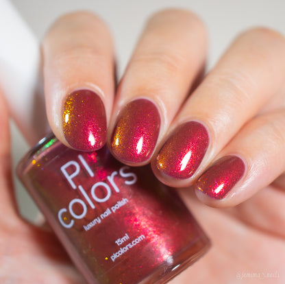 A hand with neatly manicured nails painted in Autumnal Equinox.100, a shimmering red and gold multichrome by PI Colors, holds a matching bottle labeled "PI Colors luxury nail polish.