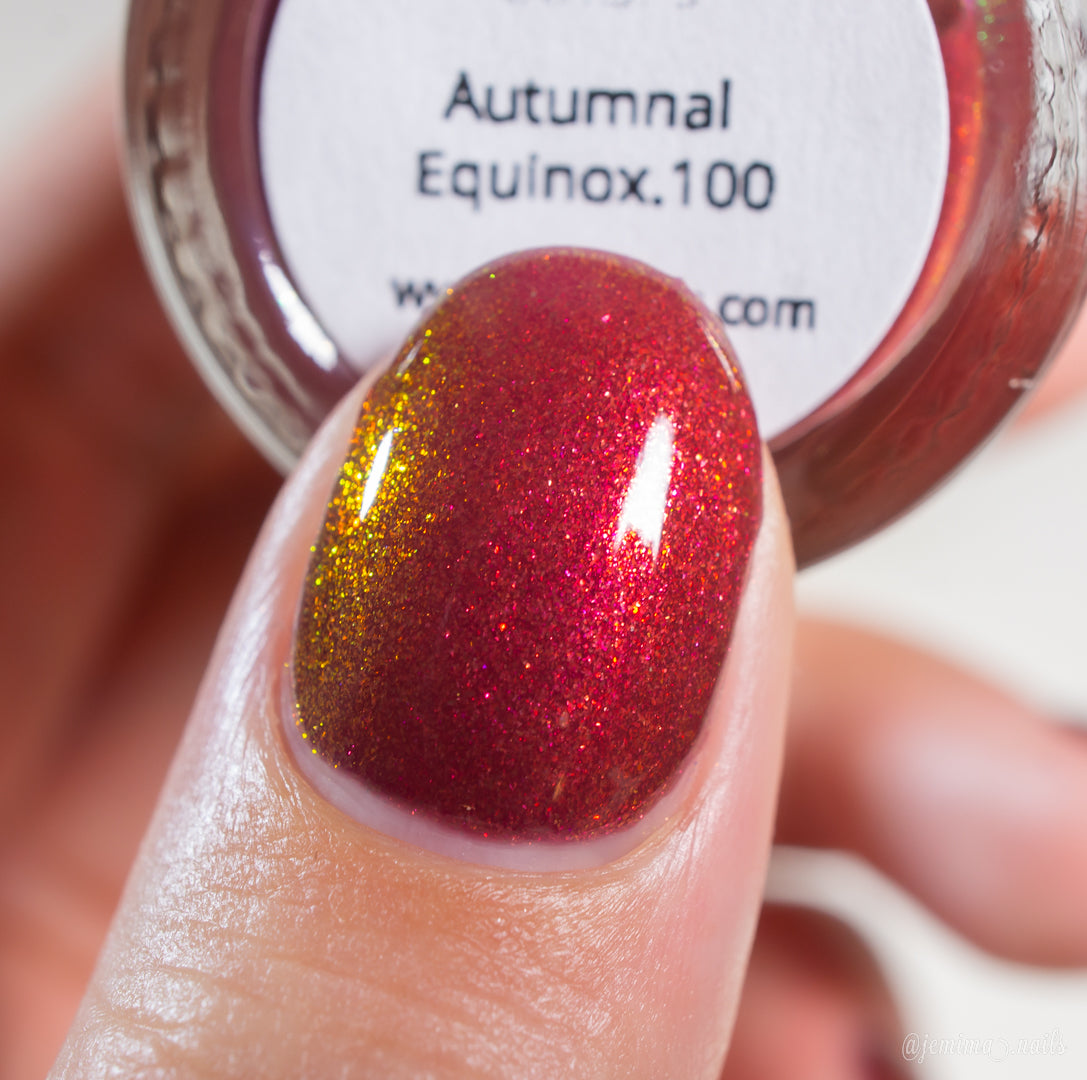 A close-up of a fingernail painted with PI Colors’ Autumnal Equinox.100, a color-shifting shimmer nail polish shifting between red, gold, and orange hues. In the background, the handmade bottle is partially visible.